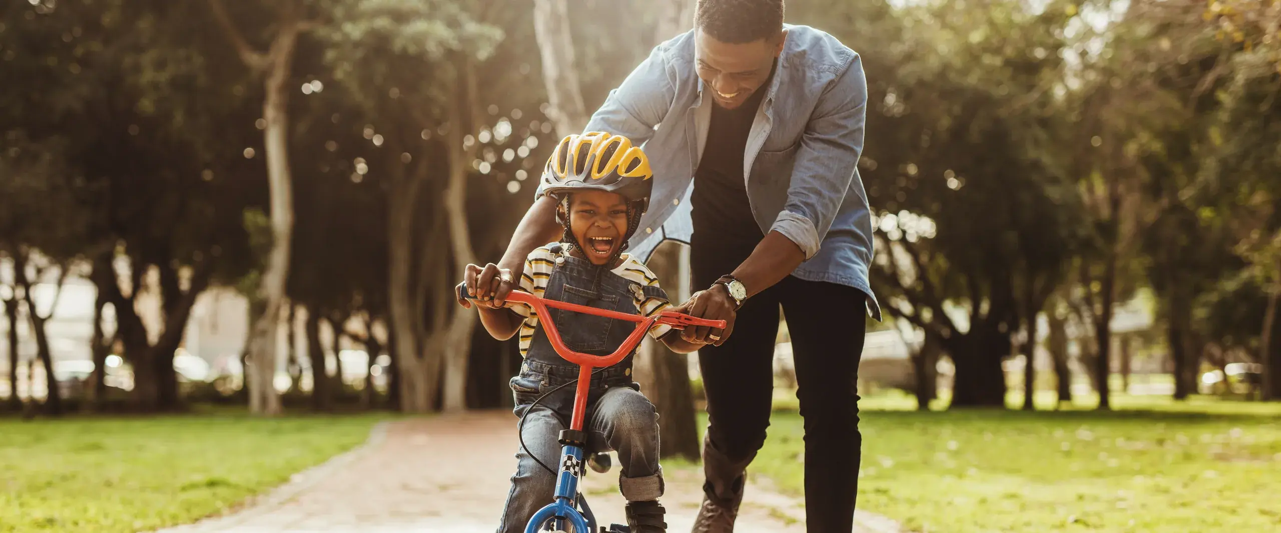 A father steadies his laughing son on a red bike along a sunlit park path.